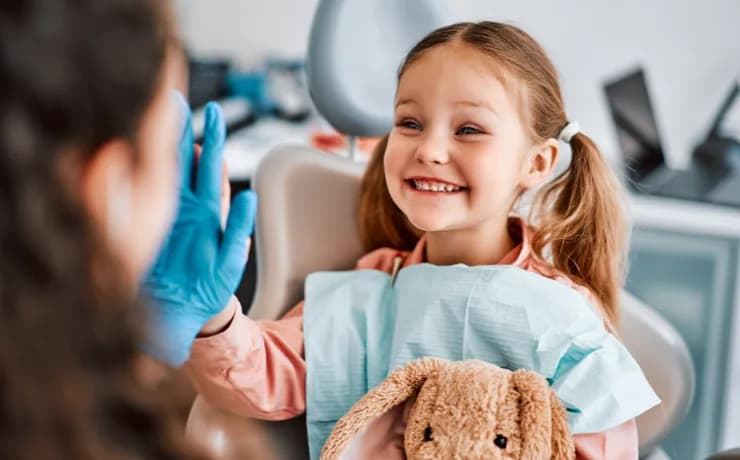 Smiling pediatric dentist comforting a young child in a colorful clinic while a relieved mom watches and checks appointments